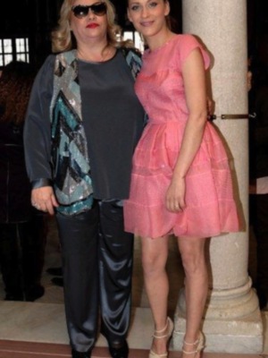 Dos personas posan juntas en una alfombra roja, uno con un vestido de lentejuelas y pantalones negros, el otro con un vestido rosa. Detalle de la alfombra roja y una columna en el fondo.