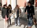 Un grupo de personas camina por una calle con edificios rojos y blancos. Una mujer en medio lleva un abrigo beige, pantalones azules y sandalias marrones. A su izquierda, una mujer en negro con un bolso blanco y tacones altos. A la derecha, una mujer en negro con un bolso marrón y zapatos marrones. Un hombre en negro con una chaqueta negra y pantalones azules.