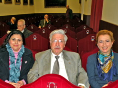 Tres personas sentadas en una sala con sillas rojas y un fondo de paredes blancas. La imagen parece ser del interior de una sala de conferencias o reuniones, con personas sentadas en filas.