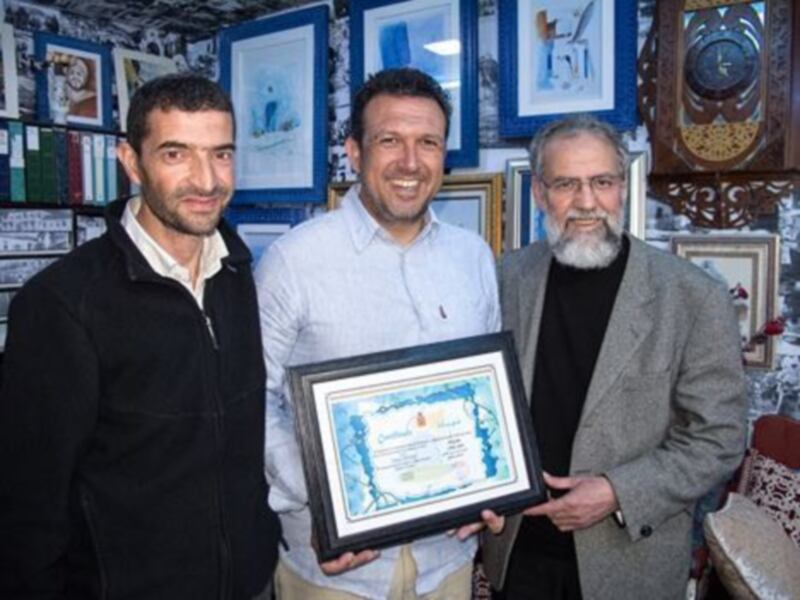 Dos hombres sonríen mientras uno sostiene un diploma. El ambiente es formal, con una pared llena de cuadros y un reloj de madera en la esquina.