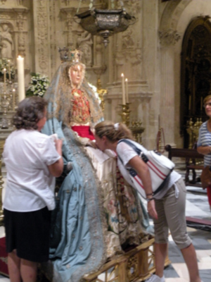 Imagen de una iglesia con una estatua de la Virgen María en el centro. Algunas personas están tocando la estatua, mientras que otras observan desde el lado. La decoración de la iglesia es elaborada con detalles en las paredes y techos.