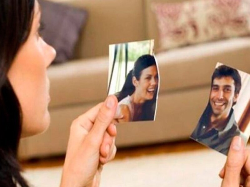 Una persona sostiene dos tarjetas de fotos, una con un hombre sonriendo y la otra con una mujer sonriente. Ambas tarjetas están colocadas de manera que parecen mirarse en el aire, creando una ilusión óptica. El fondo muestra un sofá con cojines y una pared de color beige, proporcionando contexto a la escena.