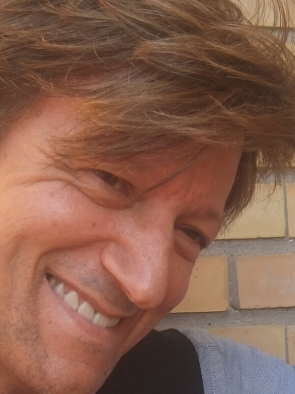 Un hombre con cabello oscuro y una sonrisa amigable, mirando directamente a la cámara. El fondo es una pared de ladrillos amarillo claro, lo que sugiere un entorno urbano.