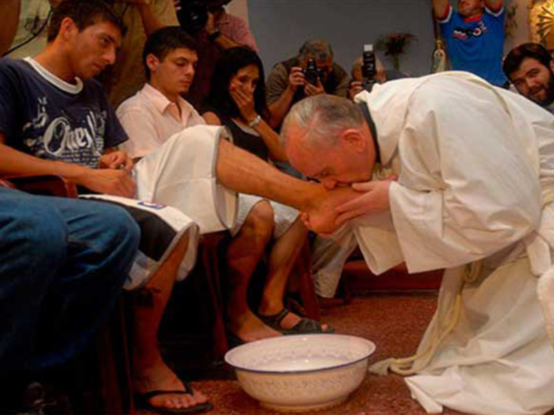 El Papa Francisco bendice a un hombre sentado en una silla blanca, mientras otros asistentes observan la ceremonia. La escena se desarrolla en un ambiente interior, con una silla y un recipiente de agua visible.