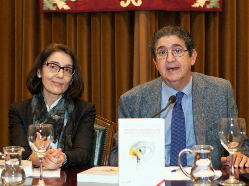 Conferencia de prensa con dos participantes sentados en una mesa con un cartel de presentación y copas de vino.