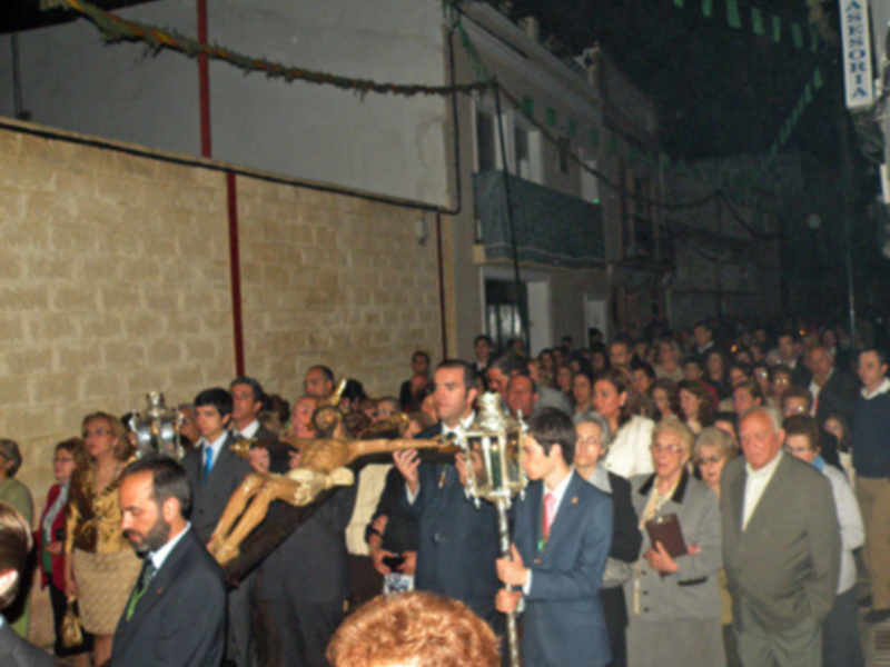 Una procesión religiosa en una ciudad, con personas vestidas formalmente y un hombre sosteniendo una cruz grande.