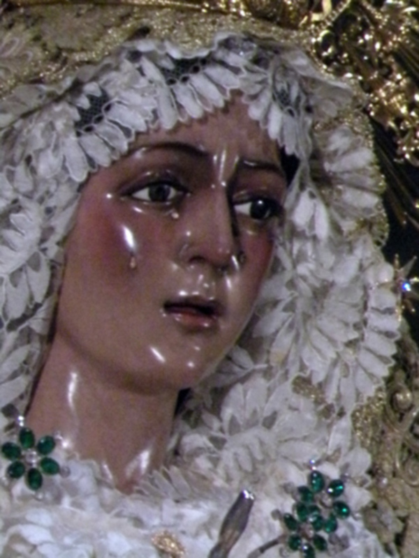 Una estatua de la Virgen María cubierta con un manto blanco y una capucha bordada. La estatua tiene ojos grandes, cejas profundas y una expresión triste. Se puede ver parte de sus ojos llorando, lo que indica una escena de dolor o sufrimiento. La estatua está adornada con joyas y broches, destacando su belleza y solemnidad.