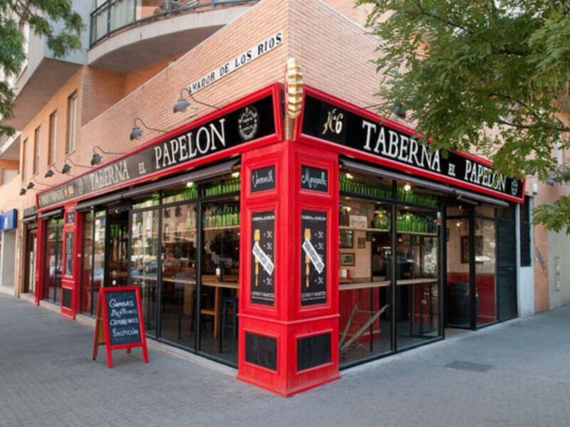 Taberna y Papeleon, ubicada en una calle con edificios de dos pisos. La fachada es roja y tiene ventanas grandes, con carteles de menús en inglés y español. Una señal de madera indica que es un restaurante. La imagen muestra una calle con árboles verdes y otros negocios en el fondo.