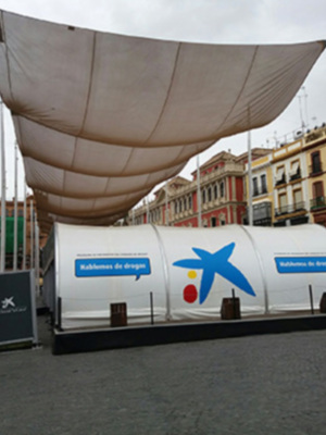 Un stand de campaña publicitario con una bandera grande y un logotipo azul y rojo. La imagen muestra edificios antiguos en el fondo, con una calle vacía y personas caminando.