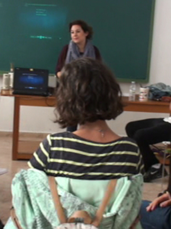 Clase en vivo con profesora explicando frente a un pizarra y proyector en un aula.