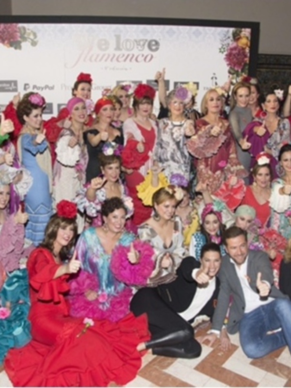 Una multitud de personas vestidas con trajes flamencos posa para una foto en un evento llamado "Flamenco Love". La imagen muestra a varias personas con diferentes estilos de trajes flamencos, desde vestidos largos hasta pantalones y blusas. La mayoría de las personas están sonriendo o haciendo gestos con los dedos, como el "OK". La escena parece ser en un lugar con decoración floral y carteles de eventos.
