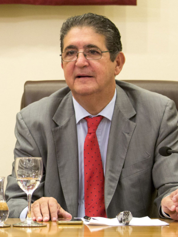 Persona en traje y corbata roja, sentado en una mesa con un micrófono y vasos, en un entorno formal.