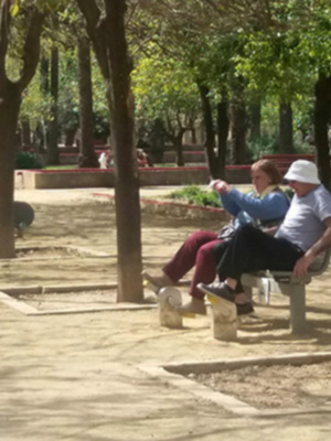 Dos personas conversan en un banco de parque, rodeadas por árboles y una zona infantil.