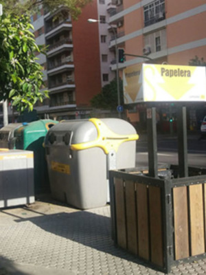 Se observan varios contenedores de basura colgados en una estructura metálica. En el fondo, se pueden ver edificios de varios pisos y un árbol en la esquina izquierda. La señalización indica que se trata de una papelería, lo cual está indicado por un letrero con una flecha amarilla y la palabra "Papelería" en letras negras.