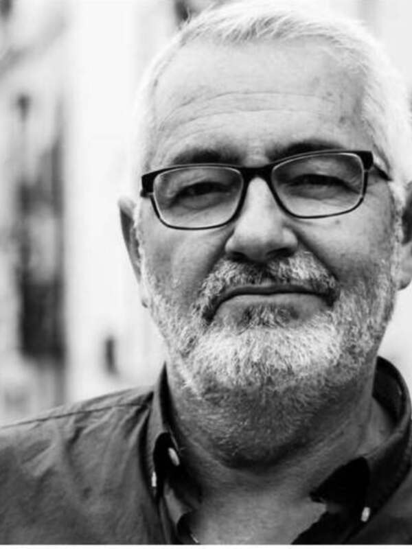 Retrato en blanco y negro de un hombre con barba y gafas, con un fondo urbano borroso.