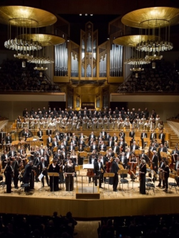 Orquesta y coro en escenario de un gran auditorio con espectadores en las gradas, bajo una impresionante estructura arquitectónica.
