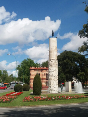 Monumento con torre y escultura en el centro, rodeado de flores y árboles. Cielo azul con nubes blancas, carros pasando en la calle.