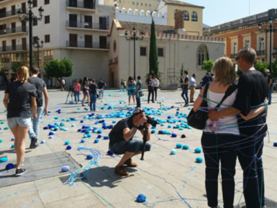 Una multitud de personas camina por un callejón con muchos globos azules sueltos en el suelo. Algunas personas están tomando fotos con cámaras, mientras otras conversan y observan el ambiente. La escena se desarrolla frente a un edificio histórico con una torre de madera en la parte superior. La gente parece estar disfrutando del día, posiblemente durante un evento o fiesta.