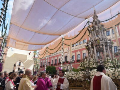 Imagen de una procesión religiosa con sacerdotes vestidos con hábitos y mantones, bajo un toldo blanco. En el fondo, edificios históricos con detalles arquitectónicos y decoración navideña.
