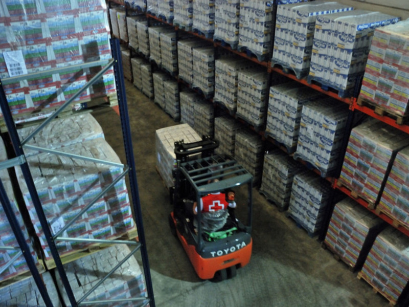 Un camión de elevación con un casco rojo y blanco está cargando cajas en una estantería de almacén. La caja que carga tiene etiquetas azules y blancas con texto en inglés. Las estanterías están llenas de cajas similares, y el suelo es de baldosas grises.