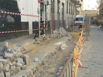 Fotografía de una calle con barricadas rojas y blancas, bloques de hormigón y una estructura metálica. El fondo muestra edificios con balcones y ventanas, algunos de los cuales tienen banderas. La imagen parece mostrar un lugar en construcción o reparación.