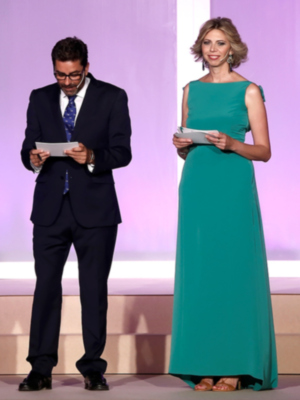 Dos presentadores en una ceremonia formal, uno vestido de negro y la otra con un vestido verde.