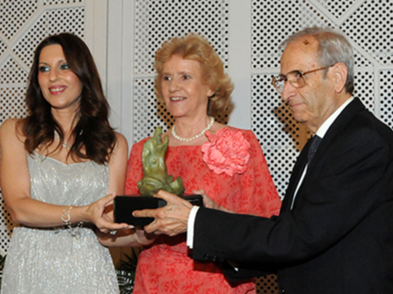 Una mujer recibe un premio de manos de una mujer mayor y un hombre mayor, todos sonrientes en un evento formal.