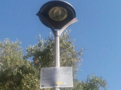 Lampara de luz solar con un cartel informativo en la base, el cielo azul y árboles de fondo.