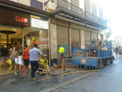 Se puede ver una calle de una ciudad con una tienda llamada "Caio" en el fondo. Hay varias personas caminando y un camión azul aparcado junto a la calle. La tienda tiene una puerta de cristal y un letrero rojo con letras grandes. La calle está pavimentada con adoquines y hay un poste de luz en la esquina.