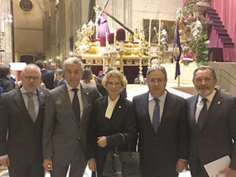 Una imagen de cuatro personas en traje formal, posando frente a una iglesia con altares y decoración. La escena sugiere un evento oficial o ceremonial.
