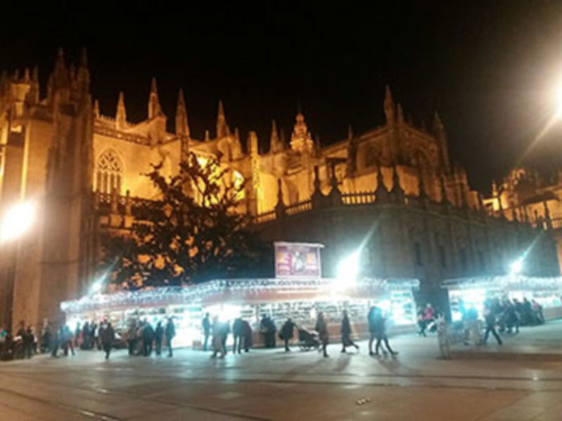 La Catedral de Sevilla iluminada por la noche, con una escena festiva en el frente. La catedral es un edificio histórico con detalles arquitectónicos góticos, y en el frente hay stands navideños iluminados con luces de colores. La plaza está llena de personas caminando y disfrutando del ambiente festivo.