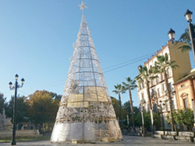 Un árbol de Navidad hecho de cristal se erige majestuosamente en el centro de una plaza, rodeado por palmeras y edificios antiguos. La estructura transparente permite ver la luz del sol filtrándose a través de sus pines, creando un ambiente festivo y soleado.