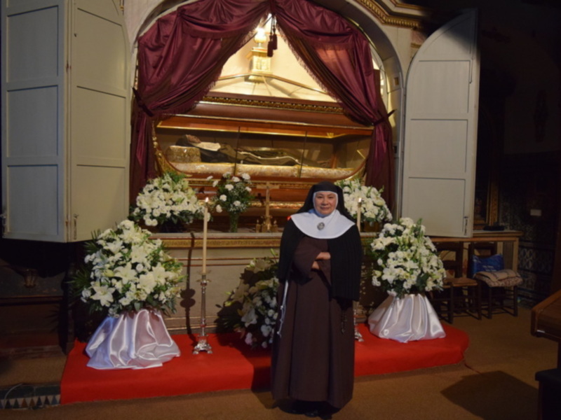 Monja en el interior de una iglesia con un ataúd y flores blancas, rodeada de decoración religiosa.