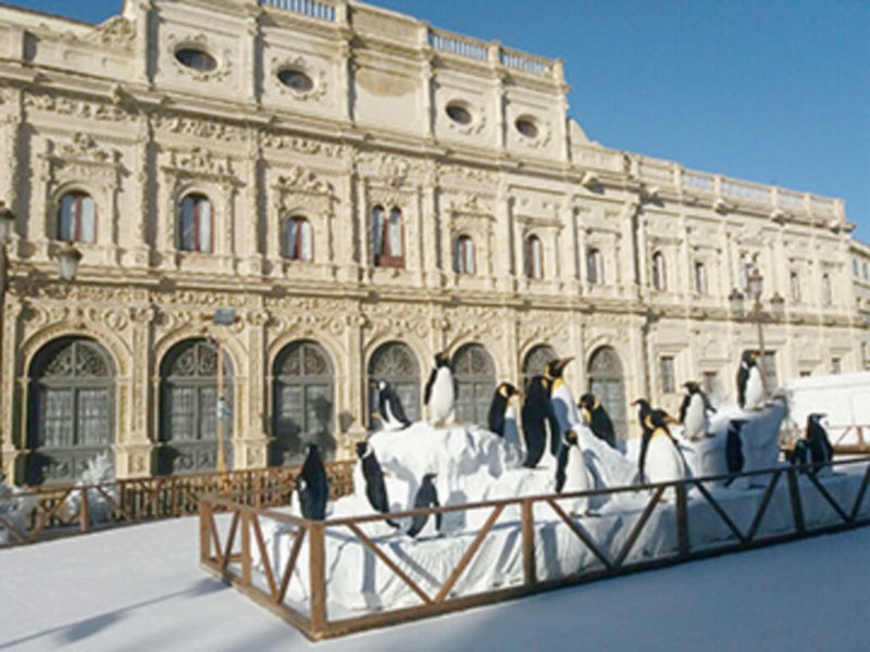 Se puede observar una escena festiva frente a un edificio histórico, posiblemente un palacio o teatro, decorado con figuras de pingüinos de hielo. El cielo es claro y el ambiente parece ser un evento o celebración navideña en una ciudad europea.