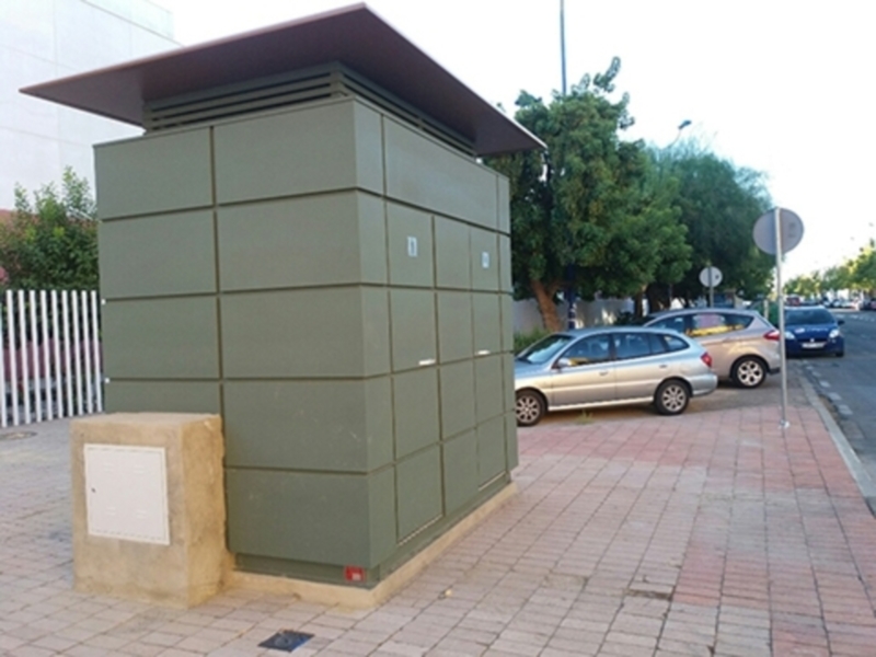 Un edificio de color verde con una cubierta roja, situado en un entorno urbano con coches estacionados y una calle a la vista.