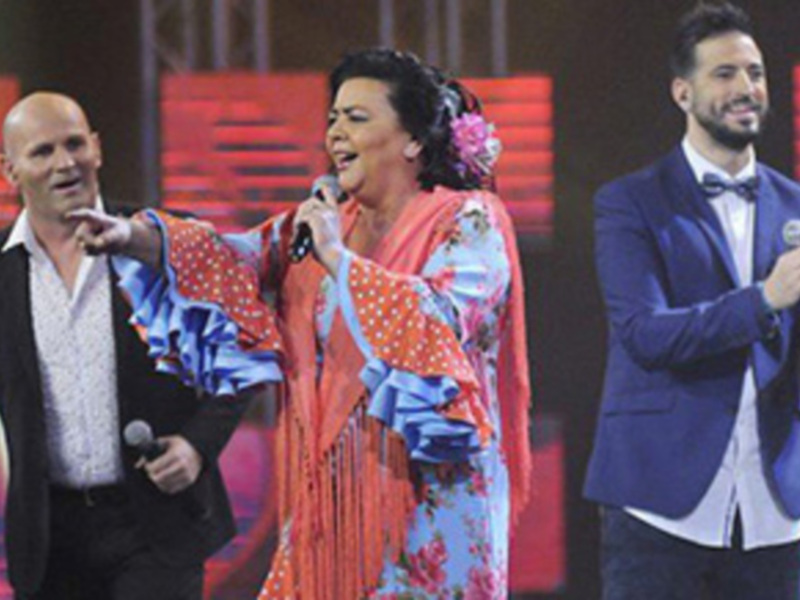 Tres personas en una escena de televisión, dos hombres y una mujer cantando. La imagen muestra a la mujer en el centro con un vestido flamenco y los hombres a sus lados, uno de ellos con una camisa blanca y el otro con un traje azul. La escena parece ser de una competencia musical o programa de entretenimiento.