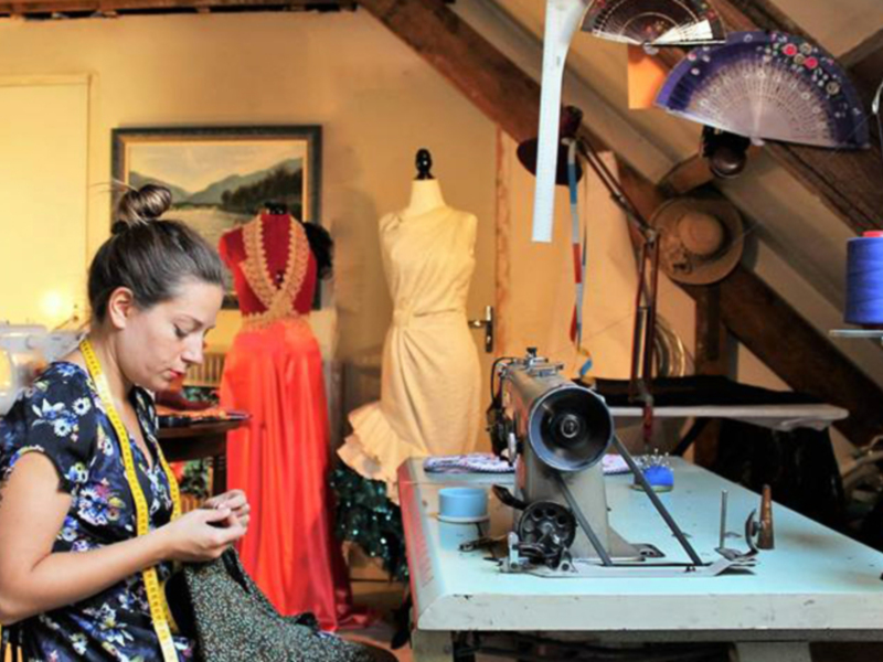 Una mujer trabaja en un taller de costura, utilizando una aguja y hilos. En la mesa hay varios accesorios de costura, incluyendo un abanico y una silla. La imagen muestra detalles de la costura, como el hilo y las herramientas necesarias para su trabajo.