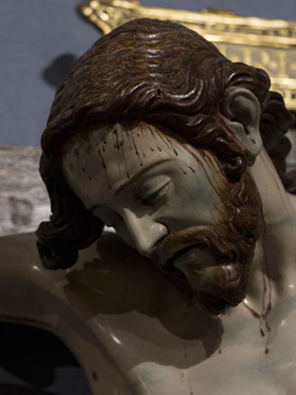 Una escultura religiosa de Jesucristo con sangre en su rostro, probablemente representando la crucifixión. La escultura está hecha de madera y tiene detalles finos en el cabello y la barba. En el fondo, se puede ver una cruz dorada que complementa el tema religioso de la imagen.