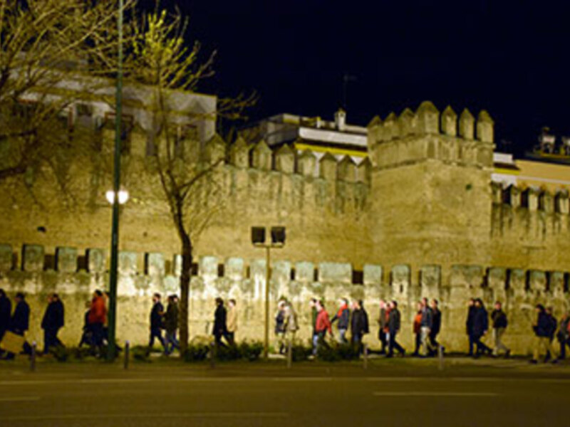 Una vista nocturna de un edificio histórico con murallas y torres, posiblemente una fortaleza o castillo. La noche ilumina el entorno con luces de calle, y en la base se pueden ver personas caminando.