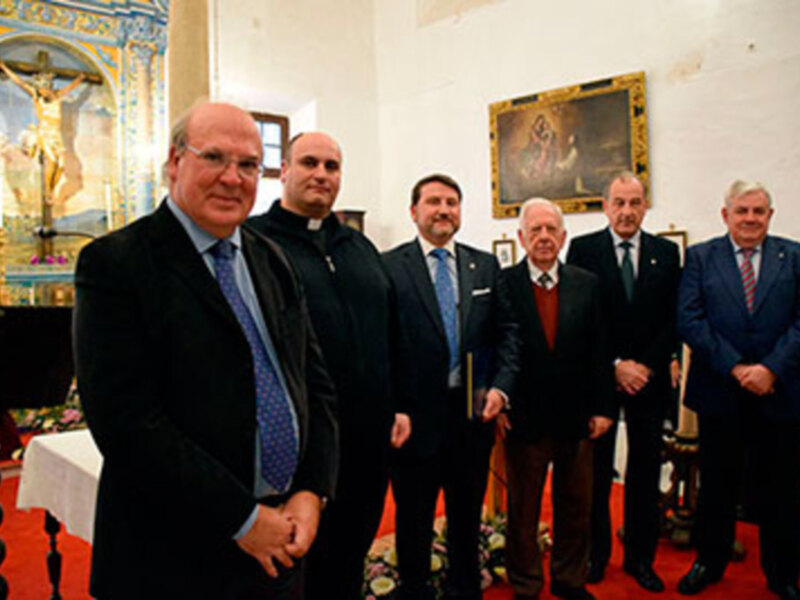 En una sala de conferencias, cinco hombres en trajes posan frente a un altar con una cruz y una imagen de la Virgen María. La escena está ambientada en un entorno histórico, con detalles arquitectónicos y decoración que sugieren una atmósfera formal y religiosa.