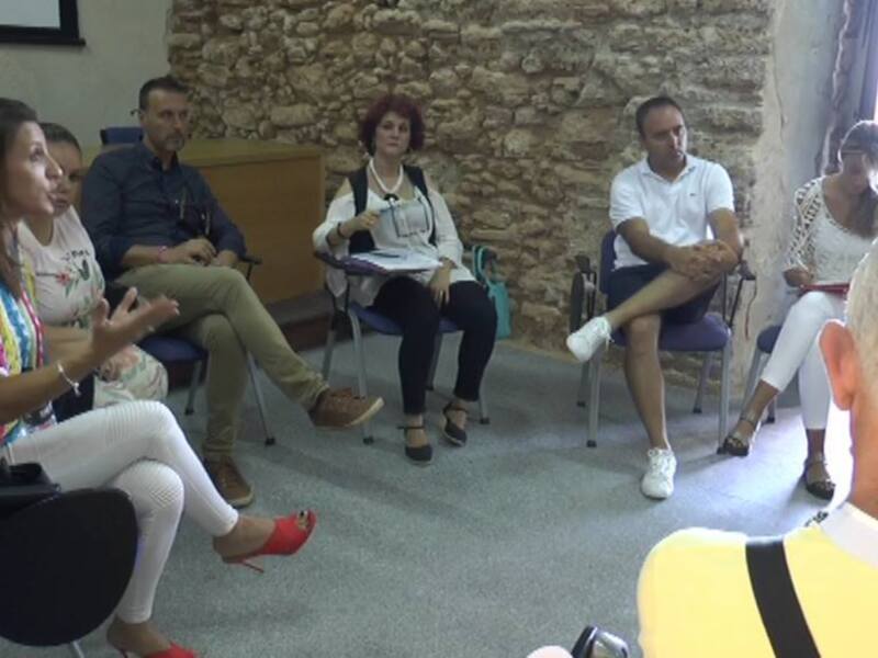 Una imagen de una sala de conferencias con personas sentadas en sillas, uno de los asistentes está hablando y las demás escuchando. La pared es de piedra, y hay un hombre en el fondo con una camiseta blanca.