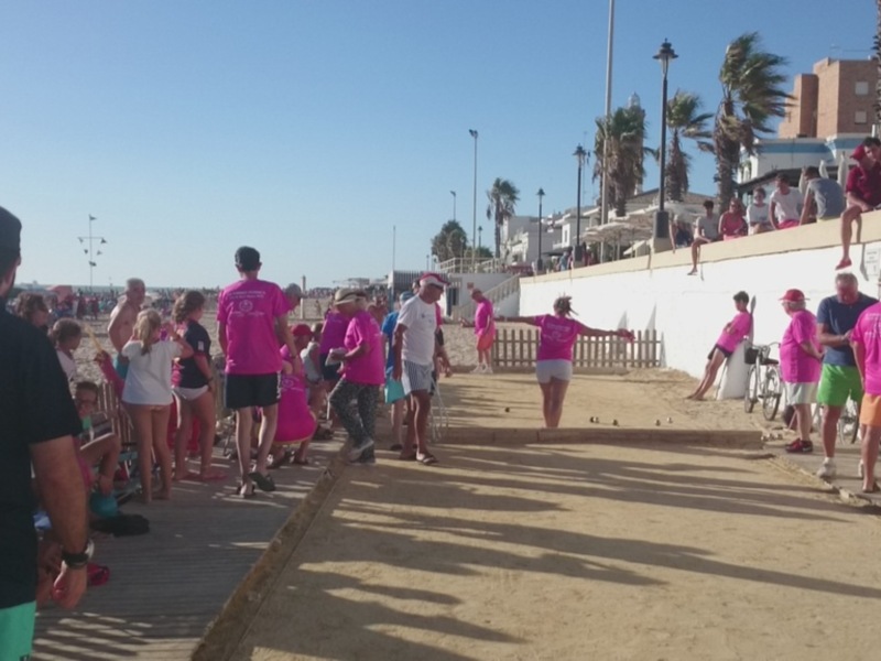 Una multitud de personas camina por una pasarela de arena hacia la playa, con palmeras y edificios en el fondo. La gente lleva ropa de playa, incluyendo camisetas rosas y pantalones cortos. El cielo es claro y azul, indicando un día soleado.