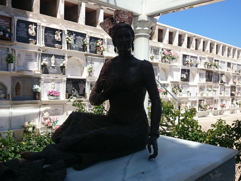 Una figura en negro se sienta sobre una lápida, rodeada de flores y monumentos funerarios en un cementerio. El cielo azul y la vegetación ajardinada completan el paisaje.