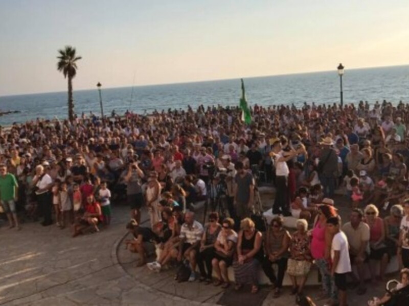 Una multitud de personas reunidas en una playa, disfrutando de la vista del mar y la música en vivo. La escena está ambientada en un día soleado, con la palmera y el agua azul como telón de fondo.