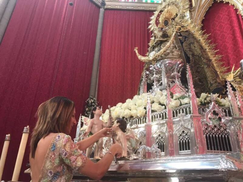 Una mujer con un vestido floral está ofreciendo flores a una estatua de cristal y oro en un altar. La imagen está ambientada en un espacio con cortinas rojas y una estructura metálica ornamental.