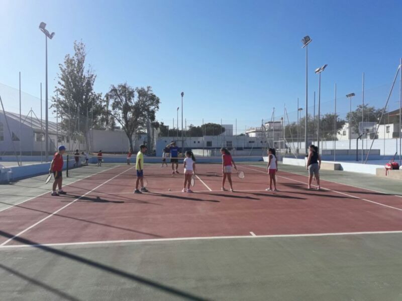 Niños jugando al tenis en una cancha de tenis al aire libre. El cielo es azul claro y hay árboles en el fondo.