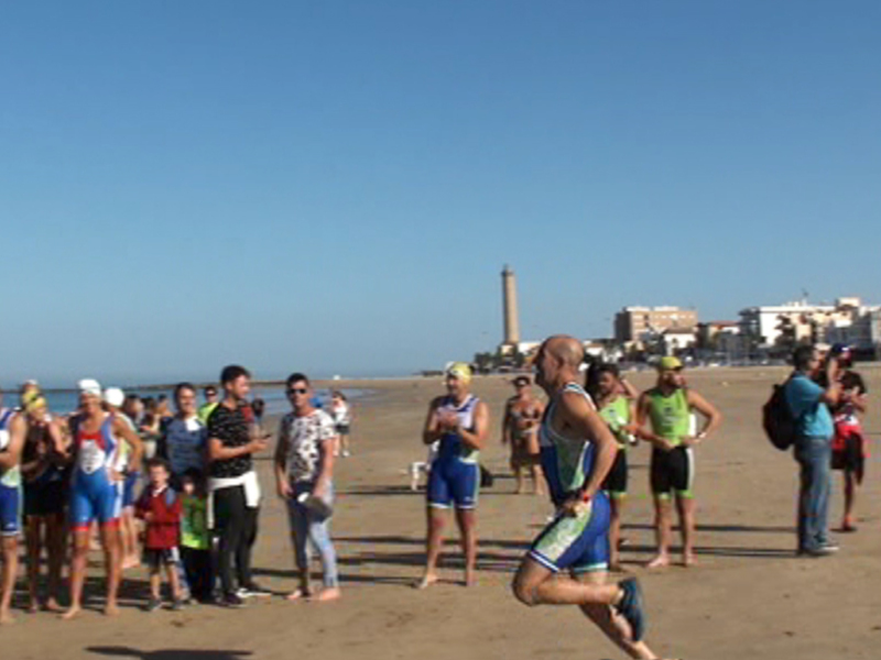 Atletas y espectadores en la línea de salida de una competición de triatlón en la playa, con el faro de Maspalomas al fondo.