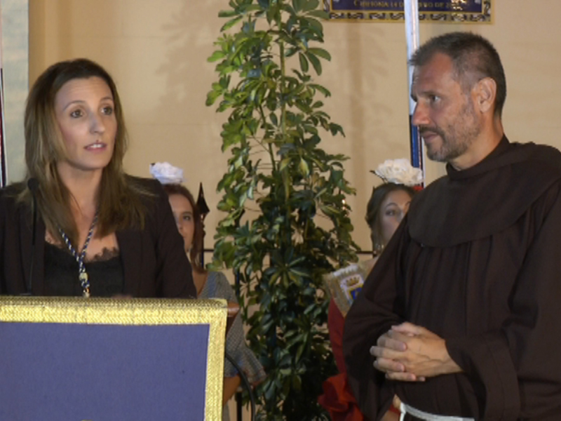 Una mujer en traje negro habla desde un estrado dorado con fondo azul. A su lado, un hombre en hábito monacal escucha atentamente. La imagen sugiere una ceremonia o conferencia formal.
