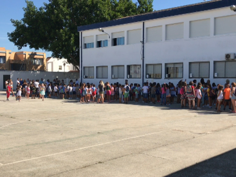 Al aire libre, una escuela con un estacionamiento de hormigón. En el fondo, hay árboles y una casa amarilla. La escuela tiene varios edificios blancos con ventanas, y en el estacionamiento hay niños de diferentes edades. La imagen muestra un día soleado con sombras en el suelo.