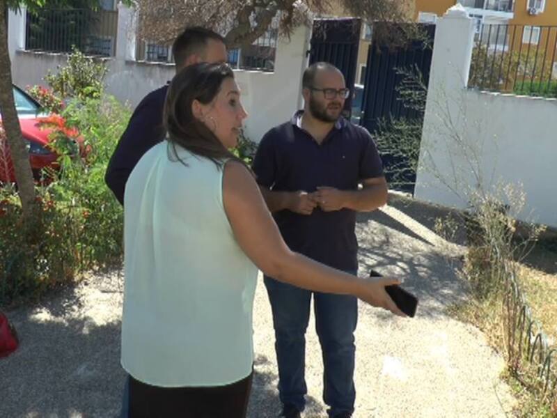 Una persona en una camiseta blanca está sosteniendo algo, mientras otra persona con un polo negro y pantalones va en su dirección. En el fondo, hay una casa amarilla con un toldo y varios coches estacionados en la calle.